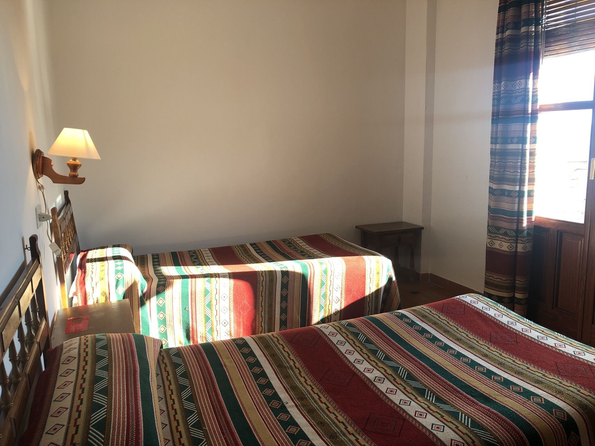 Una habitación con dos camas, cada una cubierta con una manta estampada, una mesita y una lámpara de pared.