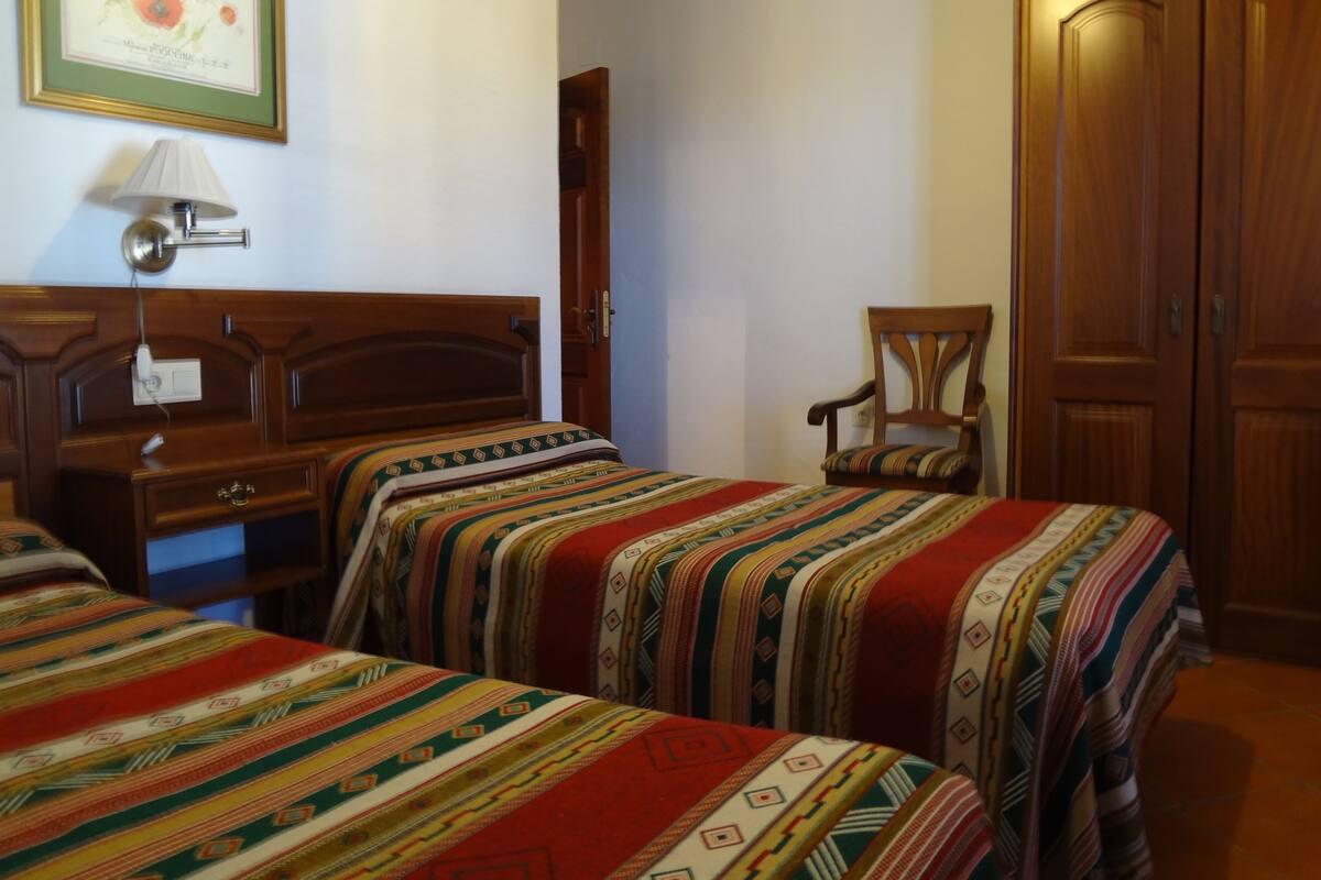 A hotel room with two beds, a wooden wardrobe, a chair, and a wall-mounted lamp.
