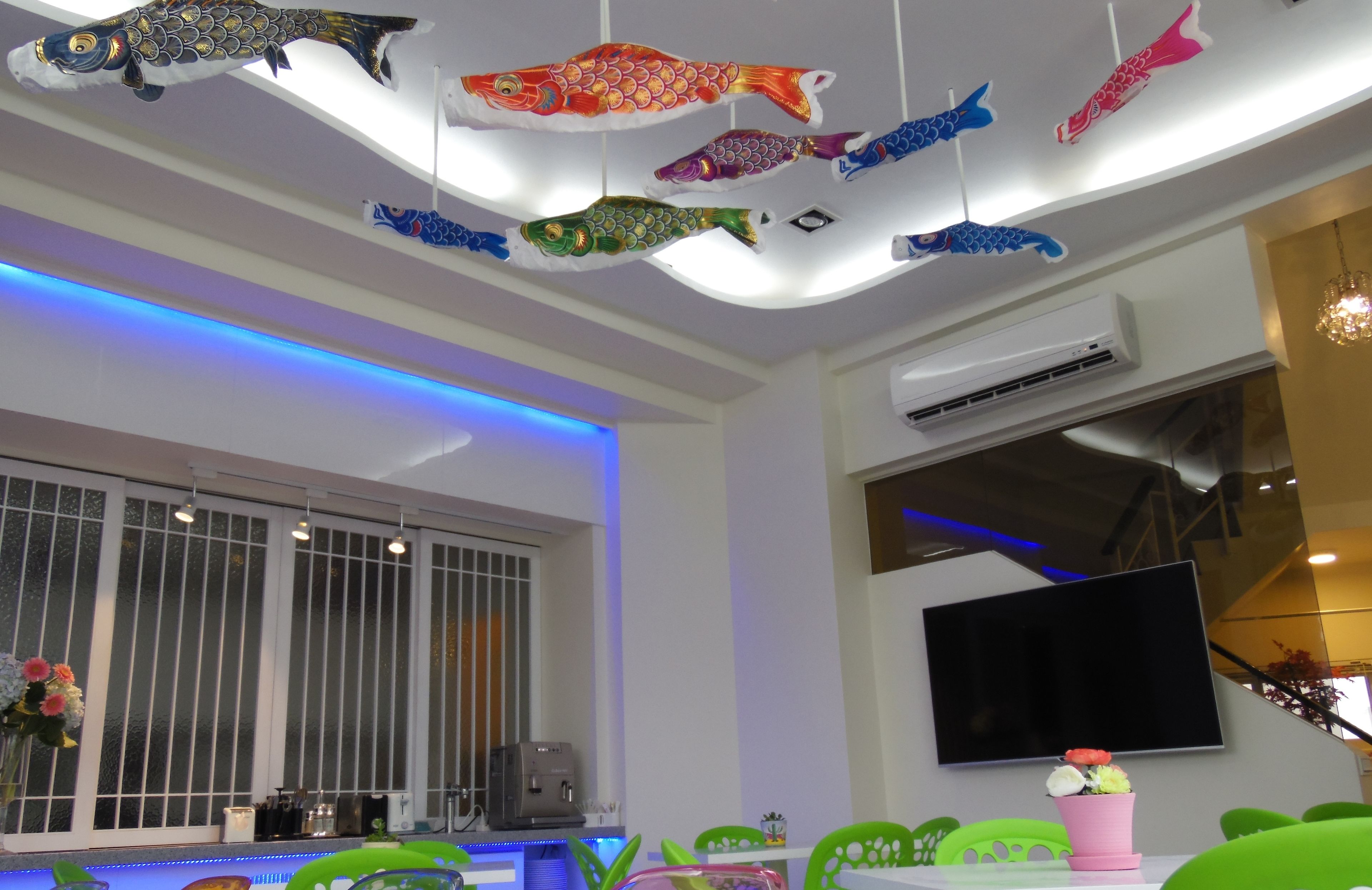 Un interior moderno con un techo decorado con coloridos adornos de peces, una ventana grande con persianas, un televisor de pantalla plana montado en la pared y una zona de comedor con sillas verdes y un jarrón de flores rosa.