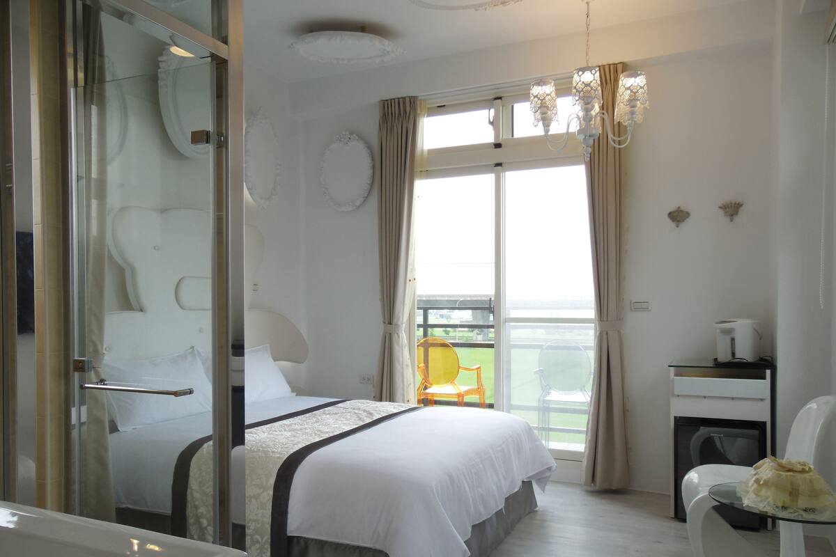 A hotel room with a bed, a desk, a chair, a balcony, and a view of the outdoors.
