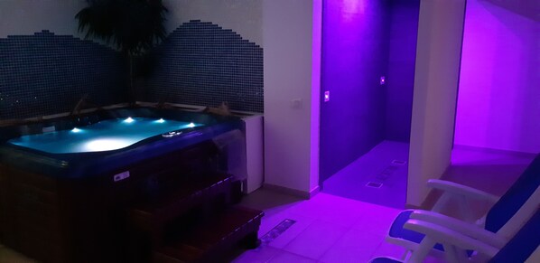 Un jacuzzi con un muro de azulejos detrás, una silla y un sendero iluminado de color morado.