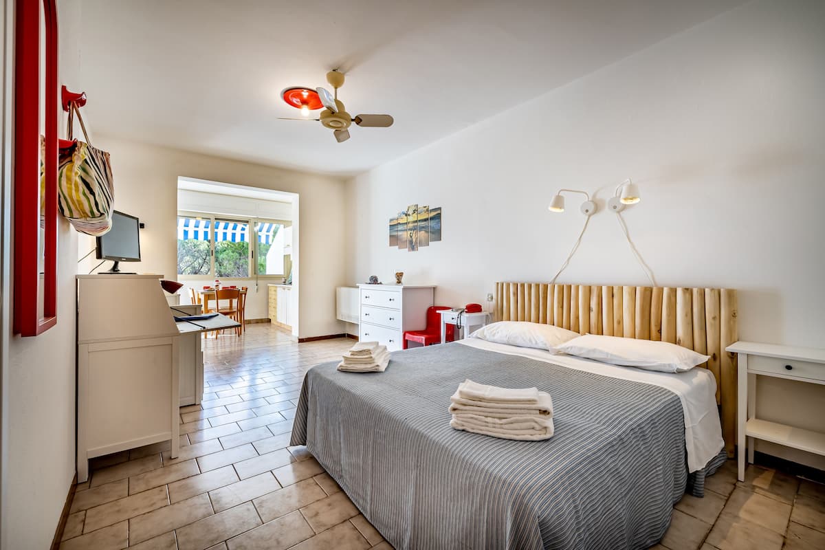 A hotel room with a bed, a desk, a chair, a wardrobe, a ceiling fan, and a window with a view of greenery.