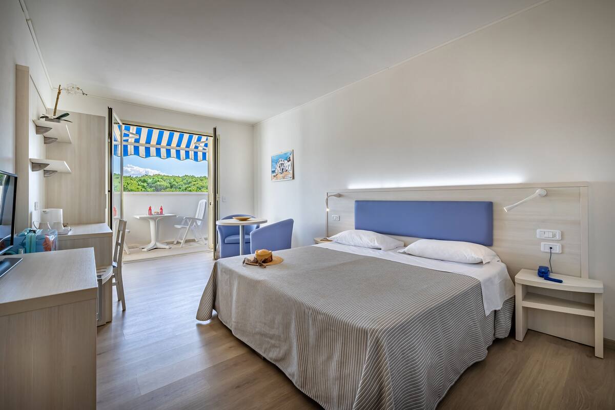 A modern hotel room with a large bed, a desk, a chair, a small table, and a balcony with a view.