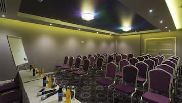 Un salón de conferencias con hileras de sillas moradas, una pizarra blanca y bebidas embotelladas sobre una mesa.