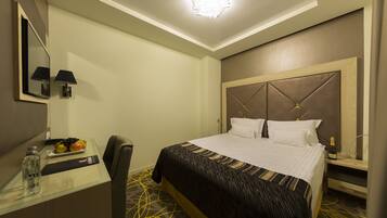 Habitación de hotel con cama, mesita de noche, lámpara y un escritorio pequeño con una botella y fruta.