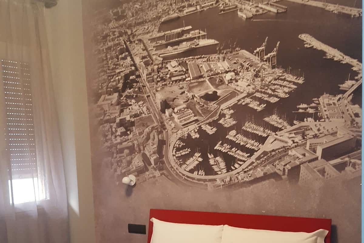 A hotel room with a bed, a large wall mural of a coastal cityscape, a window with curtains, and a red headboard.