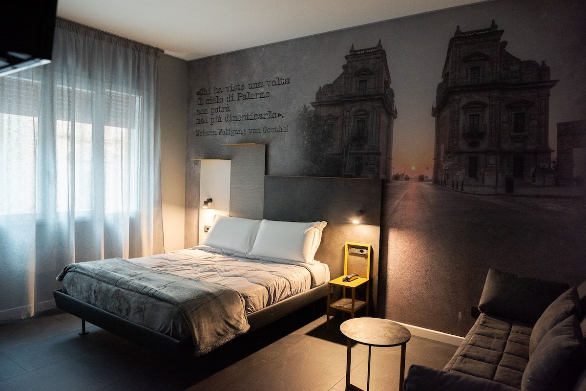 A modern hotel room with a large bed, a small round table, a sofa, and a mural of a building.