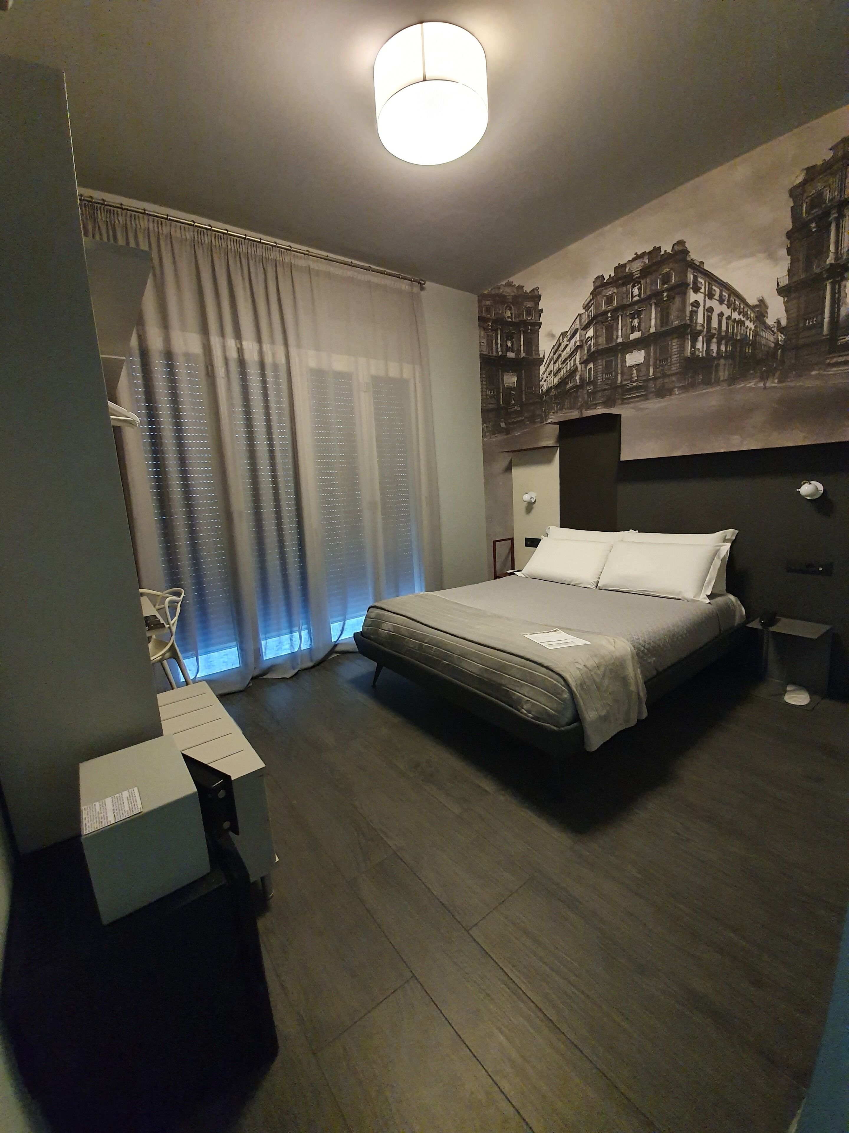 A modern hotel room with a large bed, a desk, and a window with sheer curtains.