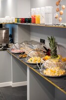 Montaje de bufé con cubiertas de vidrio transparente sobre los alimentos, que incluyen frutas y quesos, y diversas bebidas en dispensadores.