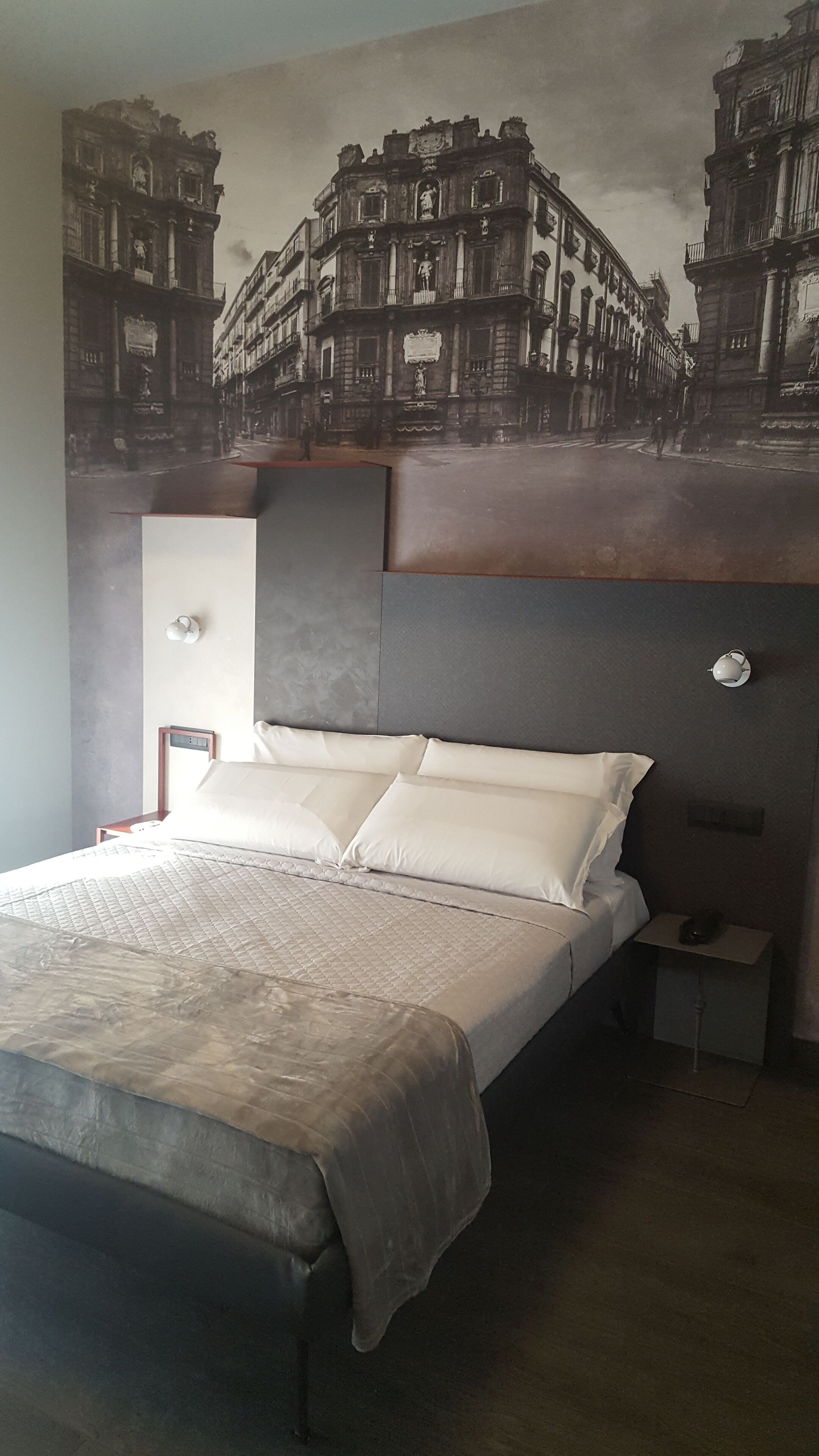 A hotel room with a large bed, a nightstand, a wall-mounted lamp, and a mural of a building.