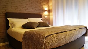 Una cama bien hecha con una manta marrón con textura, almohadas blancas y un cabecero oscuro frente a una pared de ladrillo con una lámpara de pared.