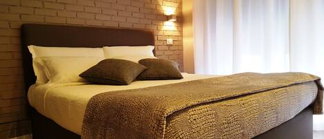 Cama bem arrumada com manta marrom texturizada, almofadas brancas e cabeceira escura em frente a parede de tijolos com luminária fixada.