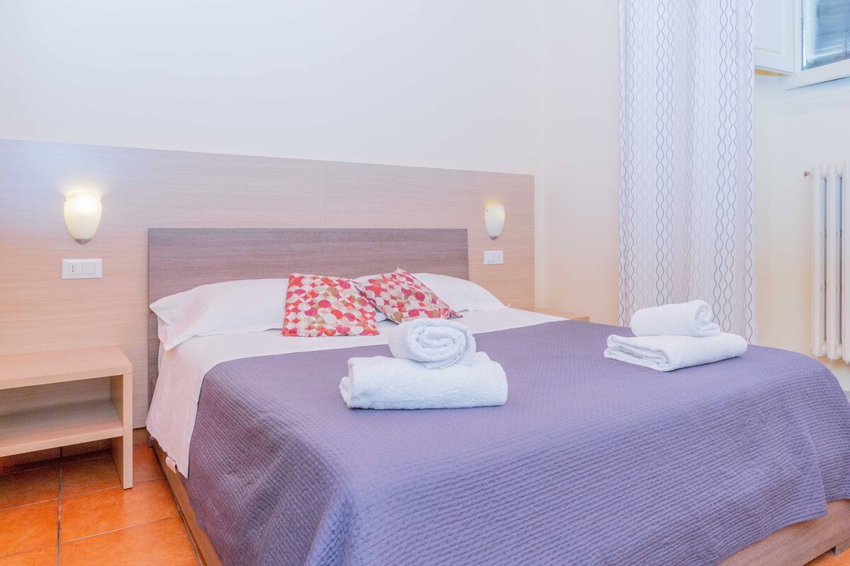 A neatly made bed with a purple bedspread, white pillows, and a rolled white towel. There are two wall-mounted light fixtures on either side of the bed.