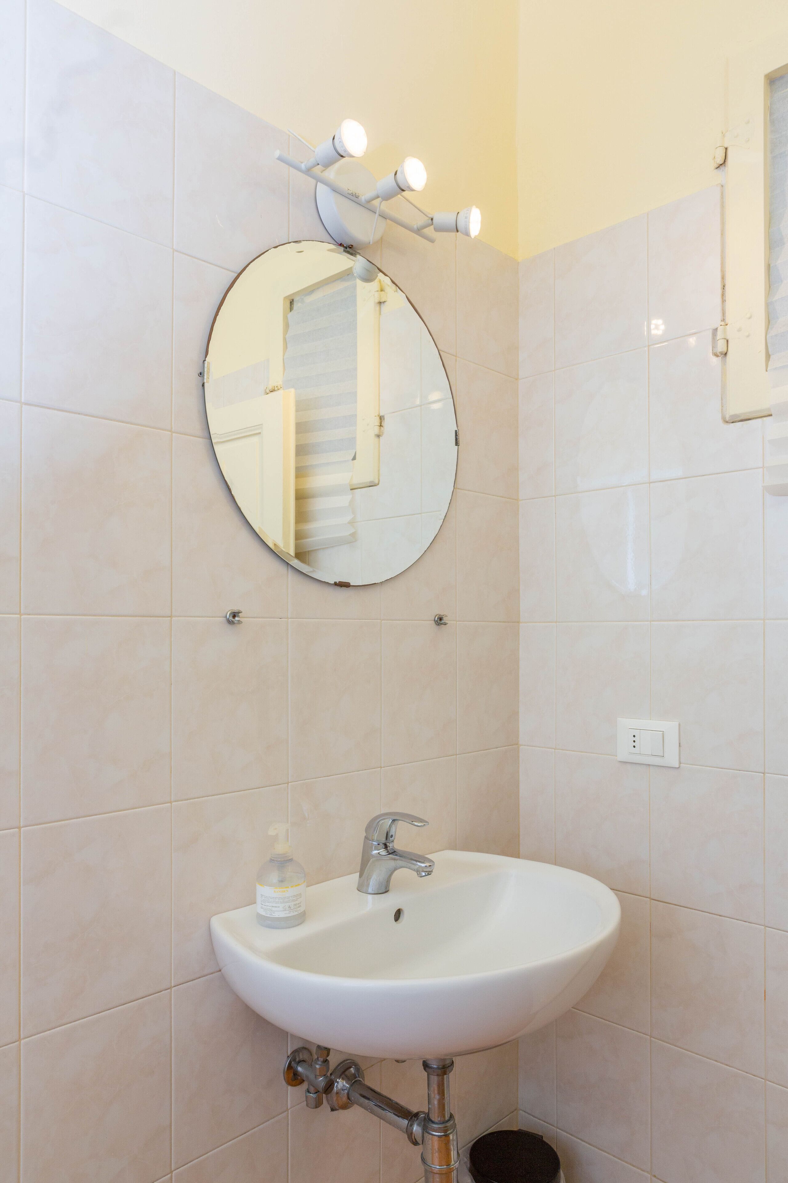 Une salle de bain avec un miroir rond, un lavabo blanc et un luminaire placé au-dessus du miroir.