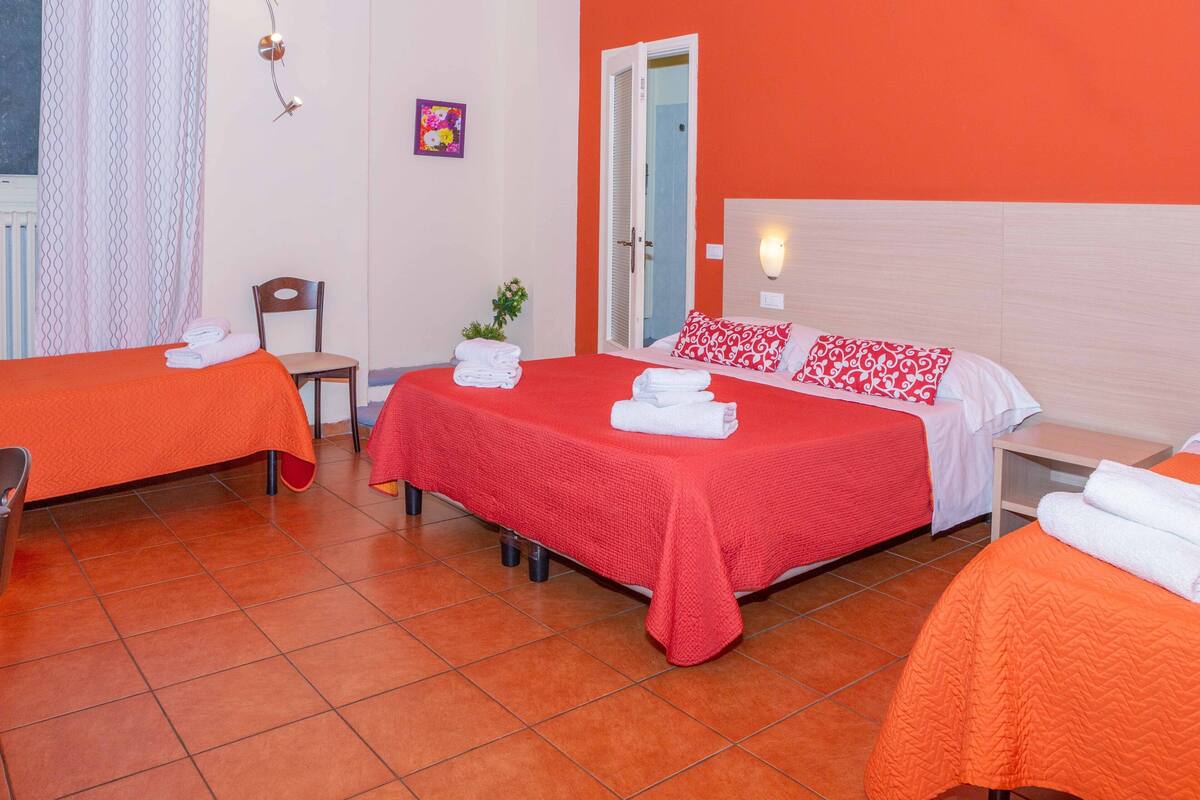 A hotel room with two beds, each with an orange bedspread and white pillows, a chair, a small table, and a wall-mounted light.