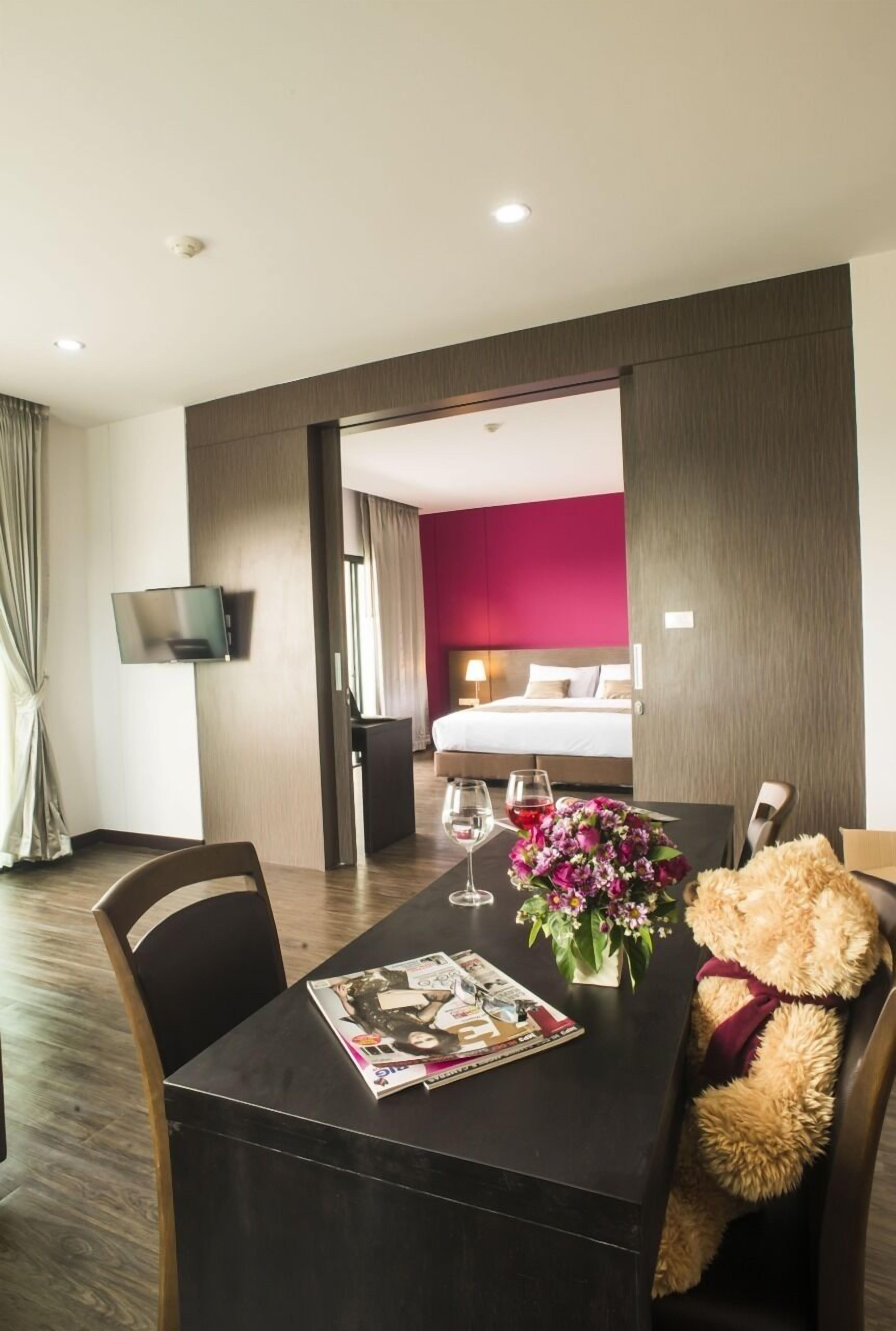 A hotel room with a dining table set with a magazine, a vase of flowers, and a teddy bear.