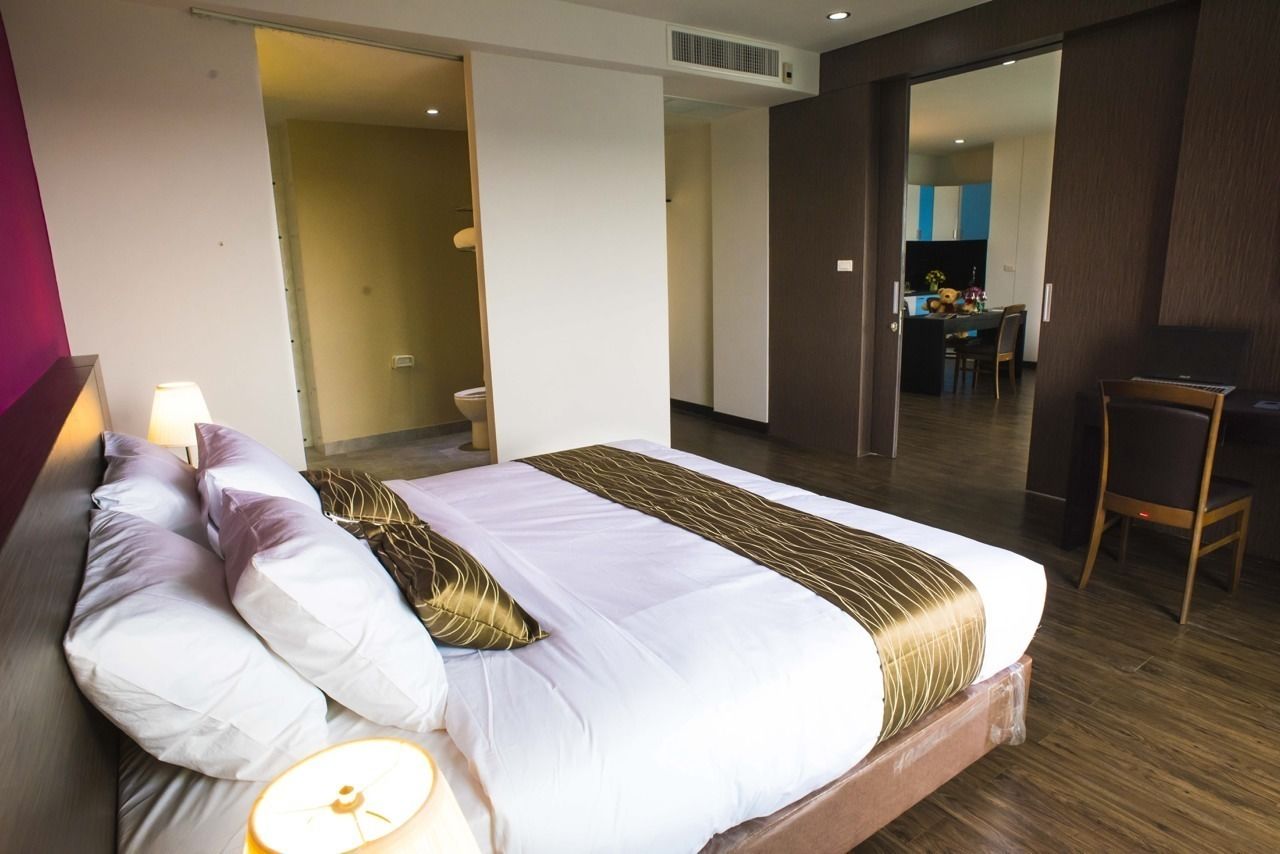 Une chambre d’hôtel moderne avec un grand lit, une lampe de chevet et une vue sur la cuisine et l’espace repas, grâce à une porte ouverte.