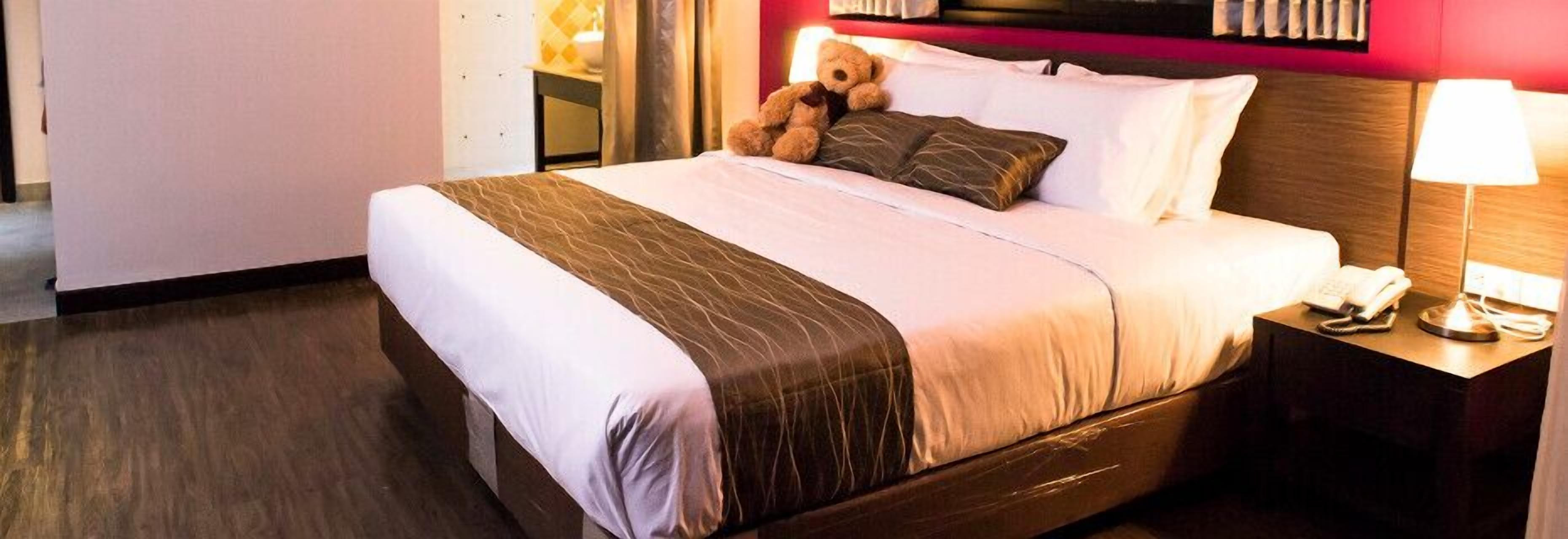 Una habitación de hotel con una cama grande, una mesita de noche con lámpara y un osito de peluche en la cama.