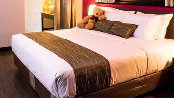 Una habitación de hotel con una cama grande, una mesita de noche con lámpara y un osito de peluche en la cama.