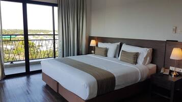 A hotel room with a large bed, two bedside lamps, a nightstand, a window with curtains, and a view of greenery outside.