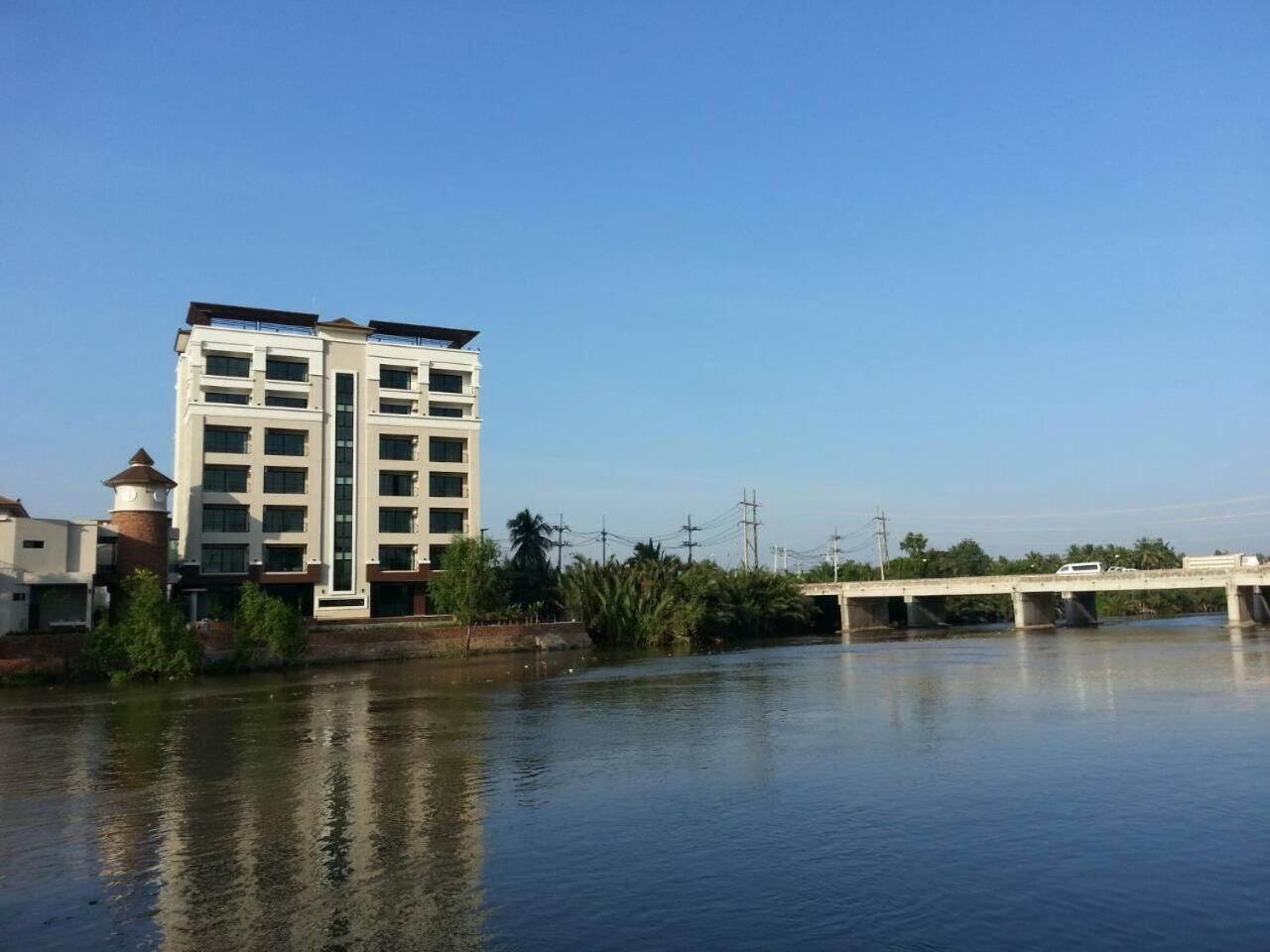 A multi-story building with a modern architectural design, situated beside a calm river with a bridge in the background.