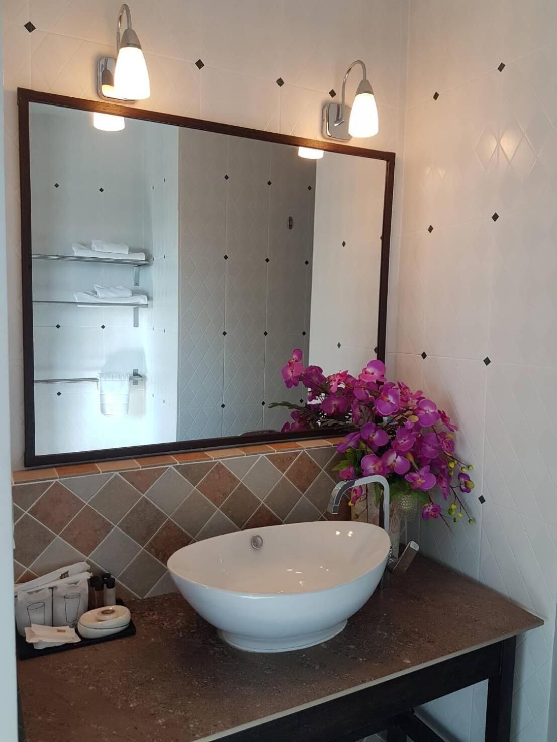 Een badkamer met een grote spiegel, een witte wastafel en een vaas met roze bloemen.