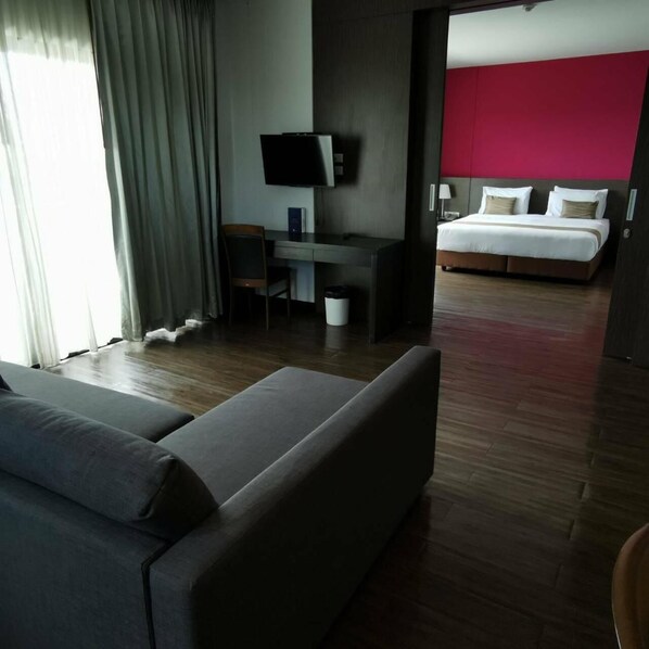 A modern hotel room with a large bed, a desk with a flat-screen TV, a sofa, and a bright red accent wall.
