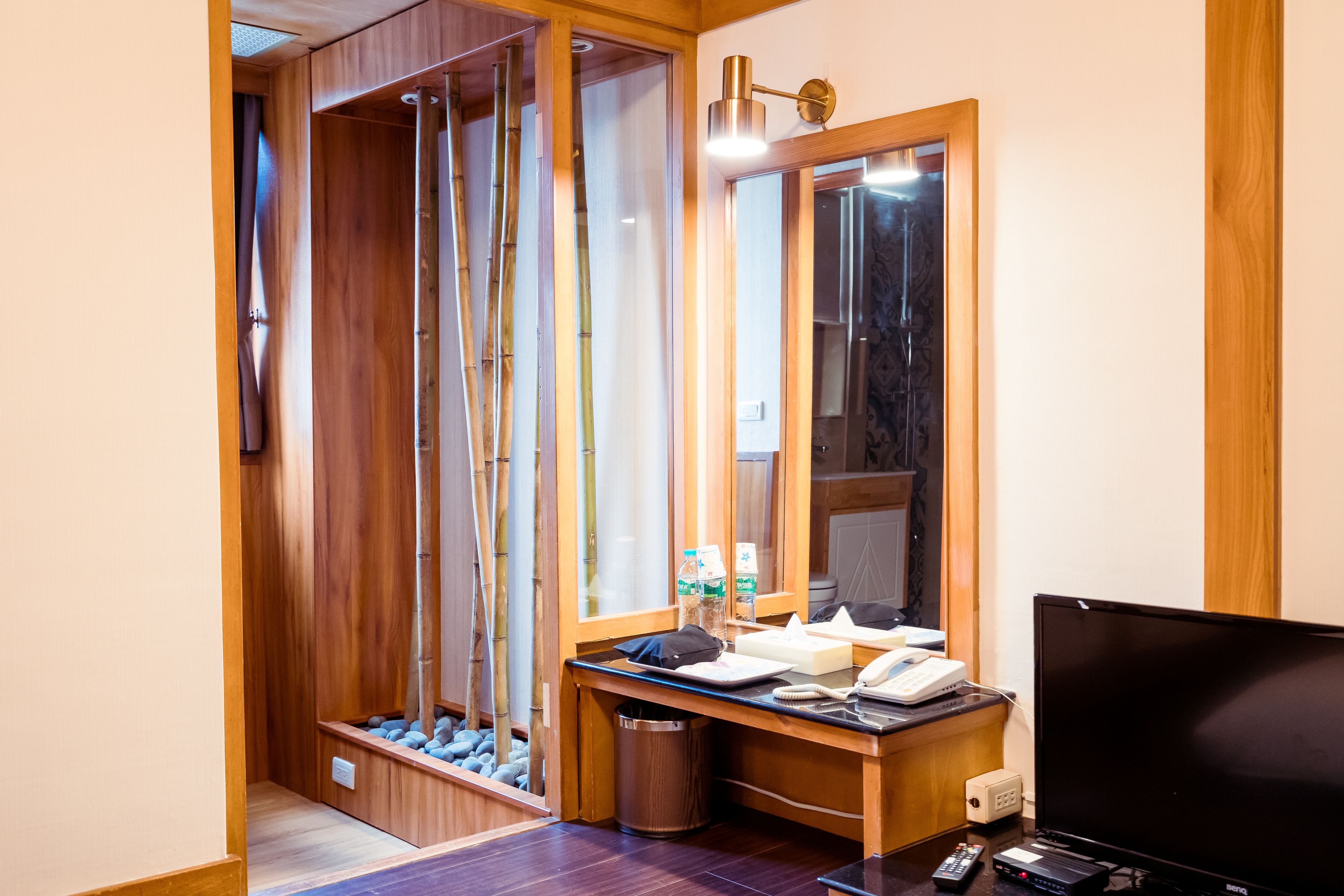 A hotel room with a wooden vanity, a mirror, a desk with a telephone and a remote control, and a television.