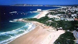 An aerial view of a coastal area with a beach, a marina with boats, and a densely built residential area with buildings and greenery. - Periwinkle Guest Lodge (Plettenberg Bay)