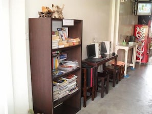 A room with a wooden bookshelf filled with books and magazines, a desk with a computer and chair, and a Coca-Cola refrigerator. - YHA Bangkok Airport - Hostel (Bang Phli)