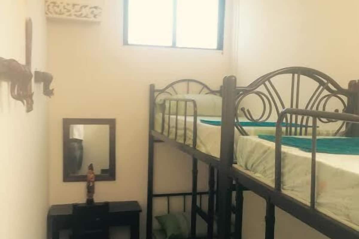 A dormitory room with bunk beds, a small desk with a mirror, and a window providing natural light.