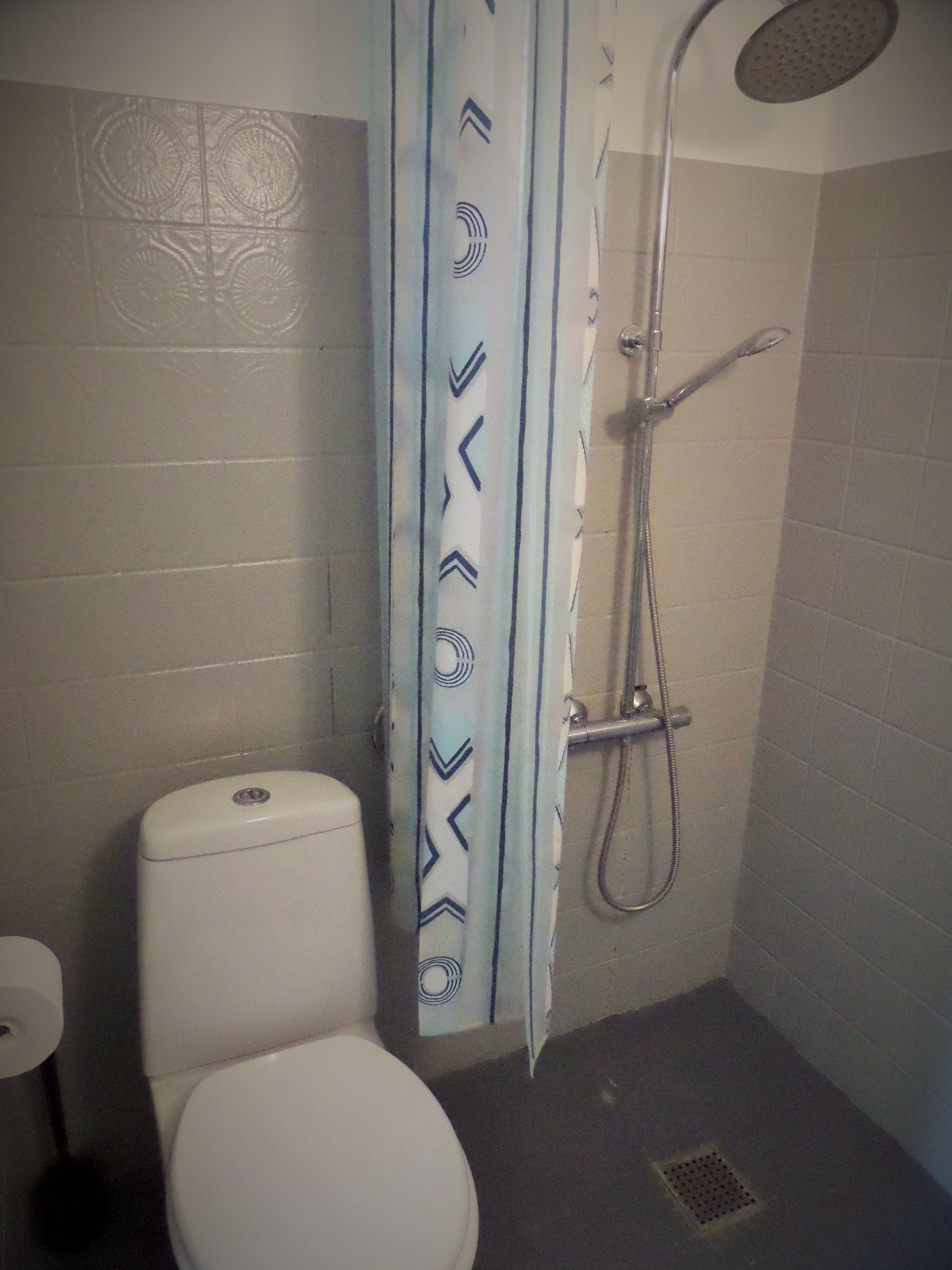Une petite salle de bain comprenant des toilettes, une douche avec un pommeau de douche amovible et un rideau orné d’un motif géométrique.