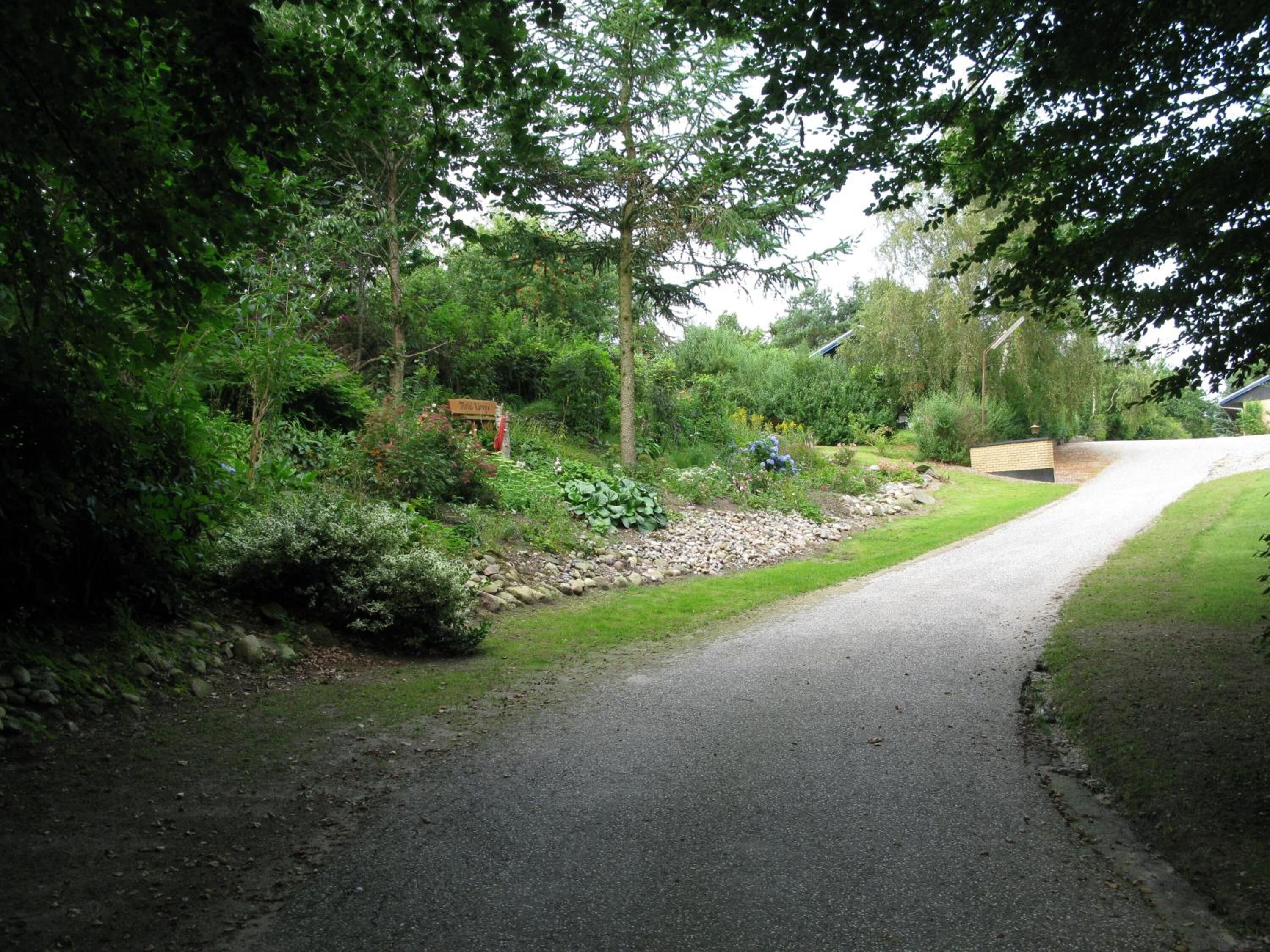 Uma estrada pavimentada serpenteia por um jardim exuberante, com uma placa de madeira e uma construção de tijolos.