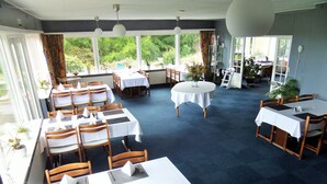 A restaurant with multiple tables and chairs, large windows with a view of greenery, and a blue carpeted floor. - Motel Europa (Svenstrup)