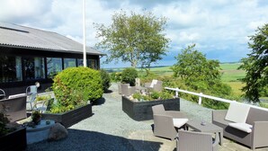 An outdoor seating area with wicker furniture and planters, a building with large windows, and a view of green fields. - Motel Europa (Svenstrup)