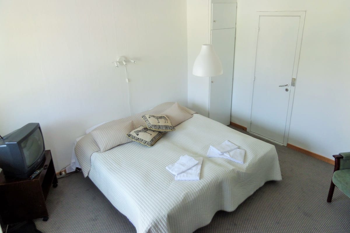 A hotel room with a bed, a television on a stand, a hanging lamp, and a door.