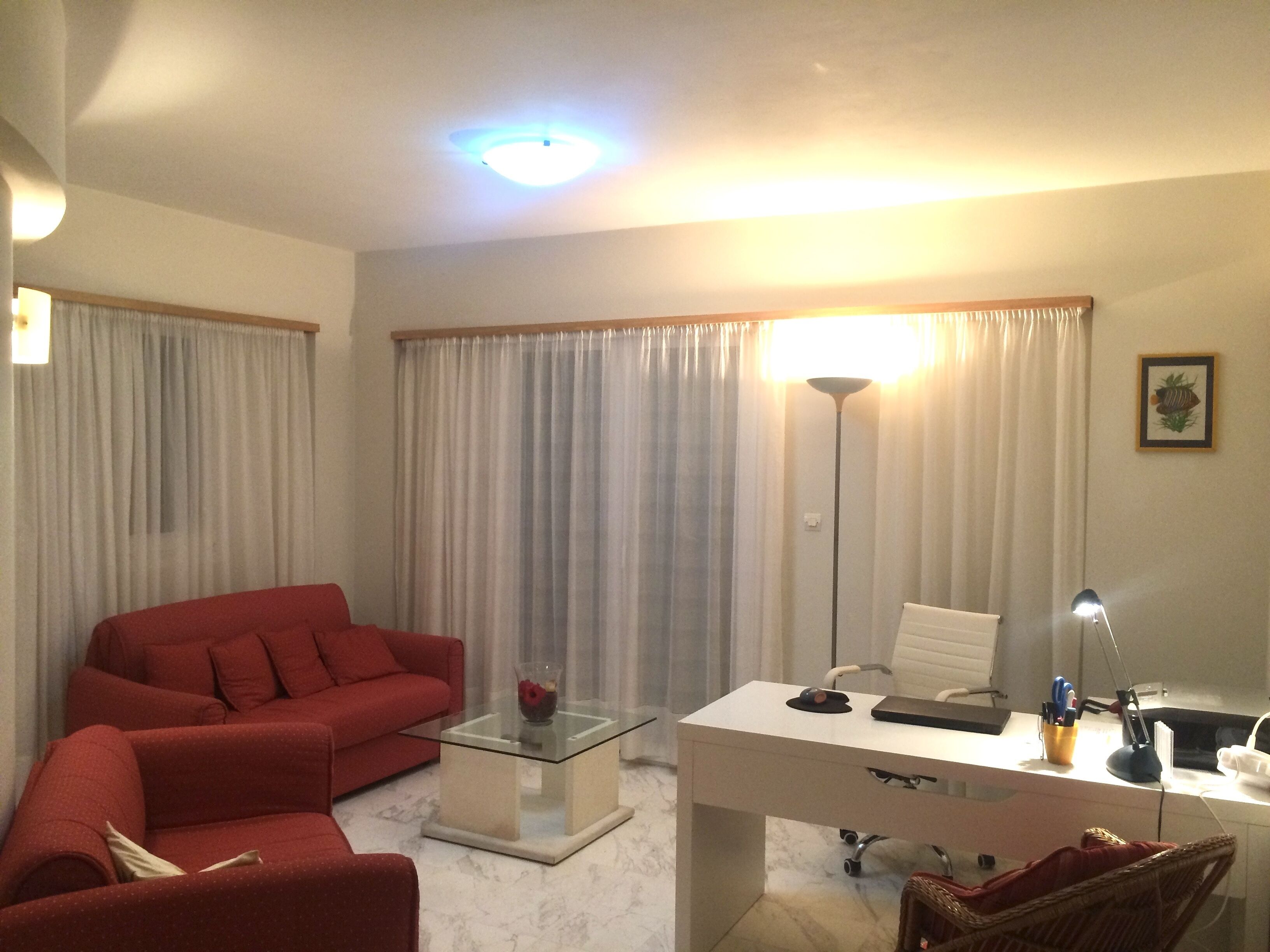 Ein Wohnzimmer mit einem roten Sofa, einem Glastisch, einem weißen Schreibtisch mit einem Computer und einer Stehlampe.