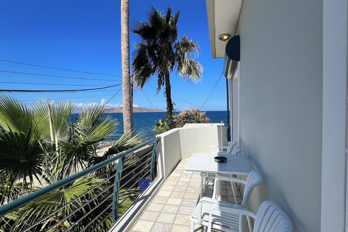 A balcony with white plastic chairs, a tiled floor, and a view of the sea and palm trees.