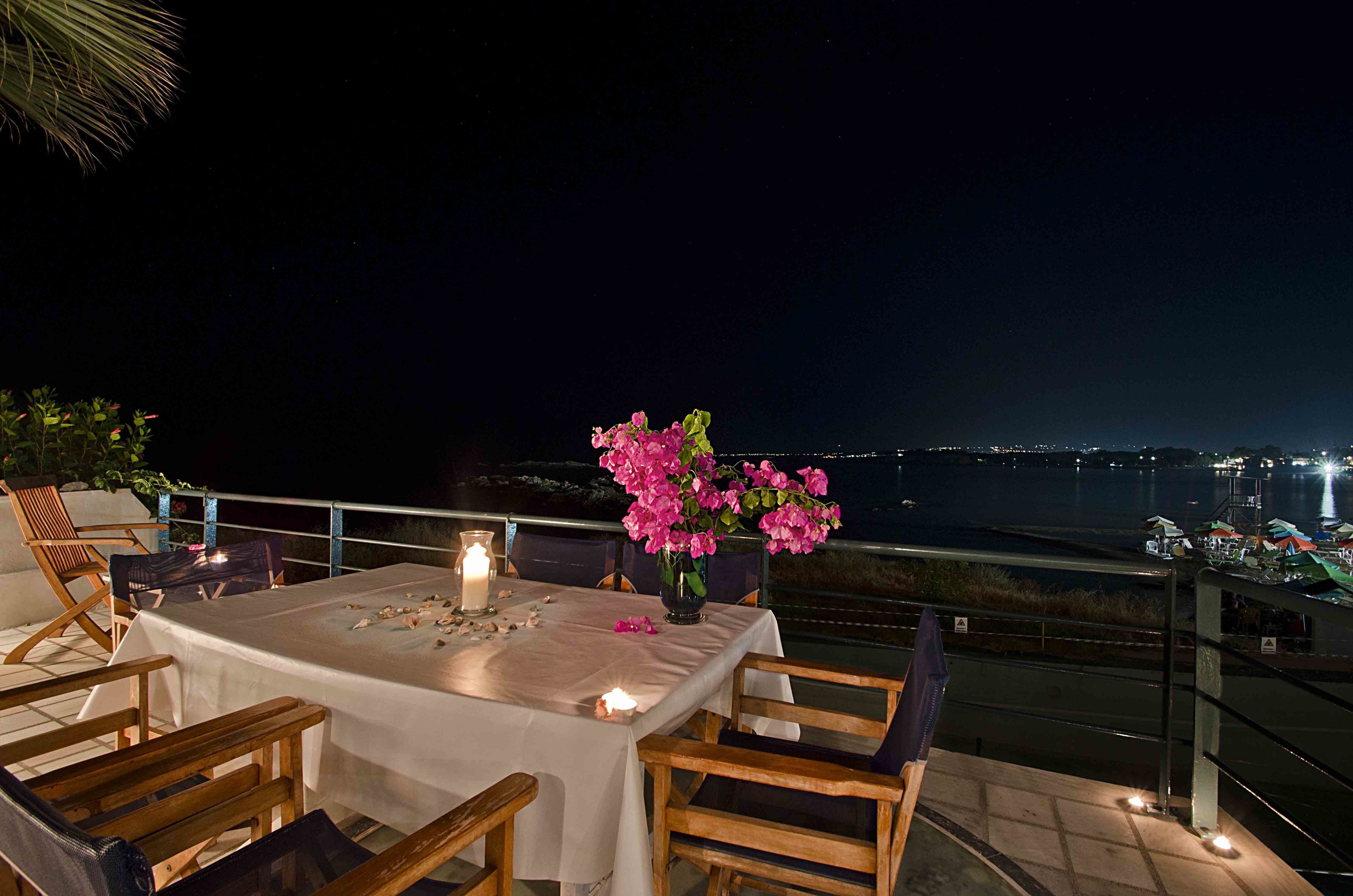 A table set for two with a white tablecloth, pink flowers, and candles on a balcony overlooking a body of water at night.
