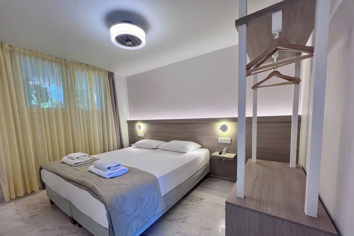 A hotel room with a bed, a nightstand, a wall-mounted light, a ceiling fan, and a wooden wardrobe.