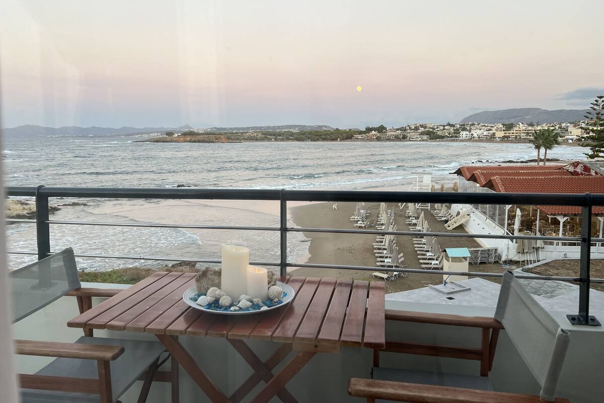 A balcony with a table, chairs, and a candle. The view includes a beach, ocean, and distant buildings.