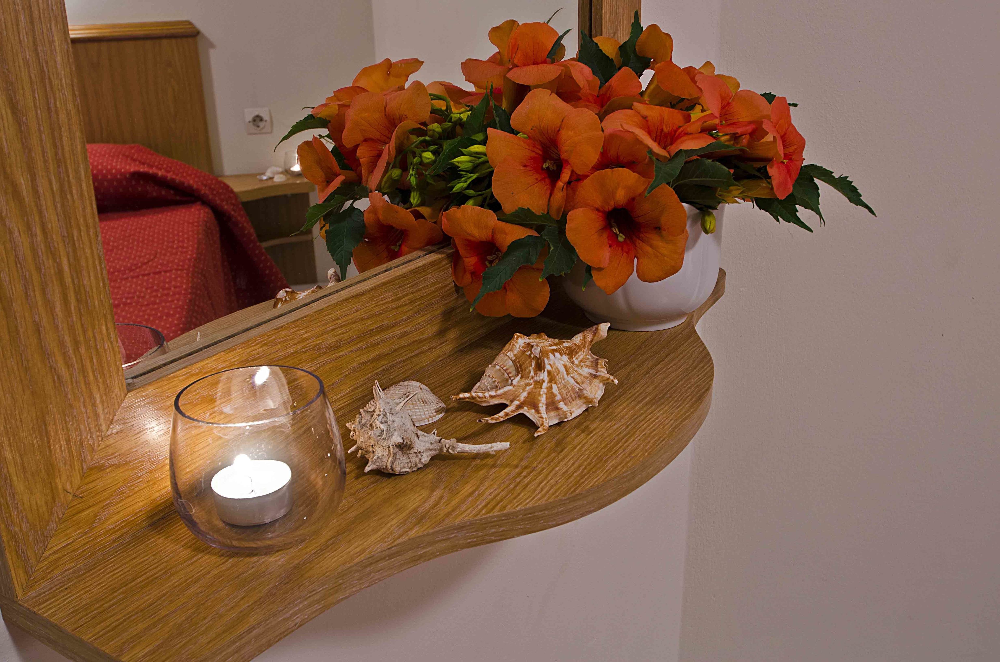 A wooden shelf with a vase of orange flowers, a lit candle, and seashells.