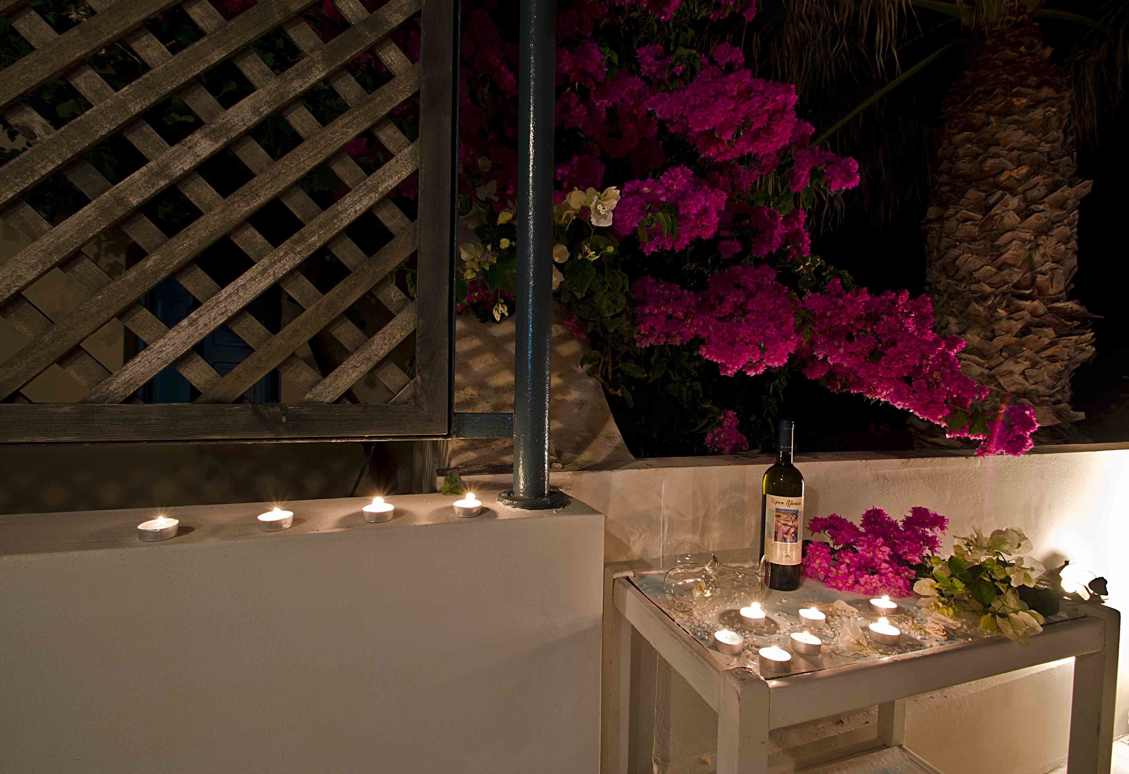 A table with candles and a bottle of wine, surrounded by pink flowers and a wooden lattice structure.