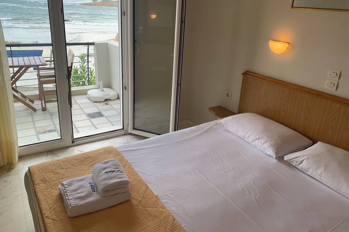 A hotel room with a bed, a small table, a chair, a view of the ocean, and a sliding glass door.