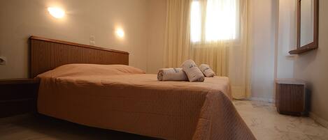 A neatly made bed with a headboard, a bedside table, and a window with curtains.