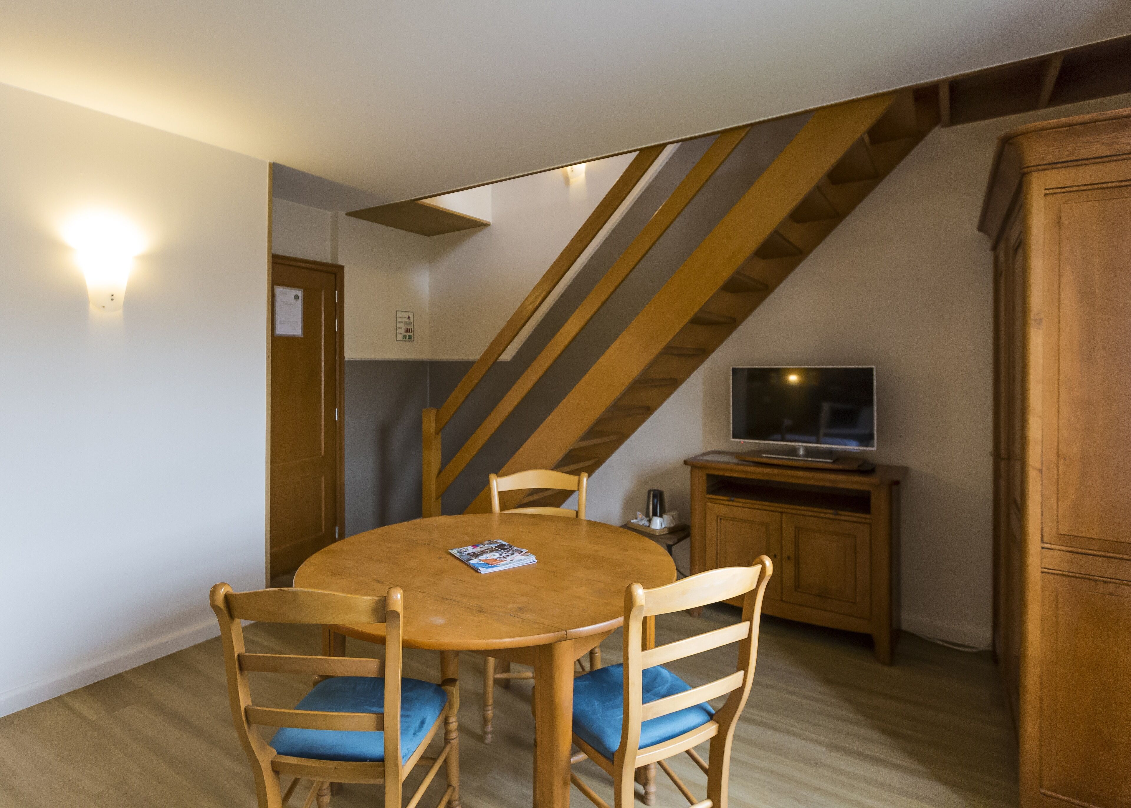 A dining area with a round wooden table and four chairs, a television on a wooden cabinet, and a staircase with wooden beams.