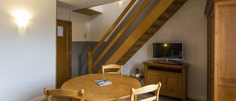 A dining area with a round wooden table and four chairs, a television on a wooden cabinet, and a staircase with wooden beams.