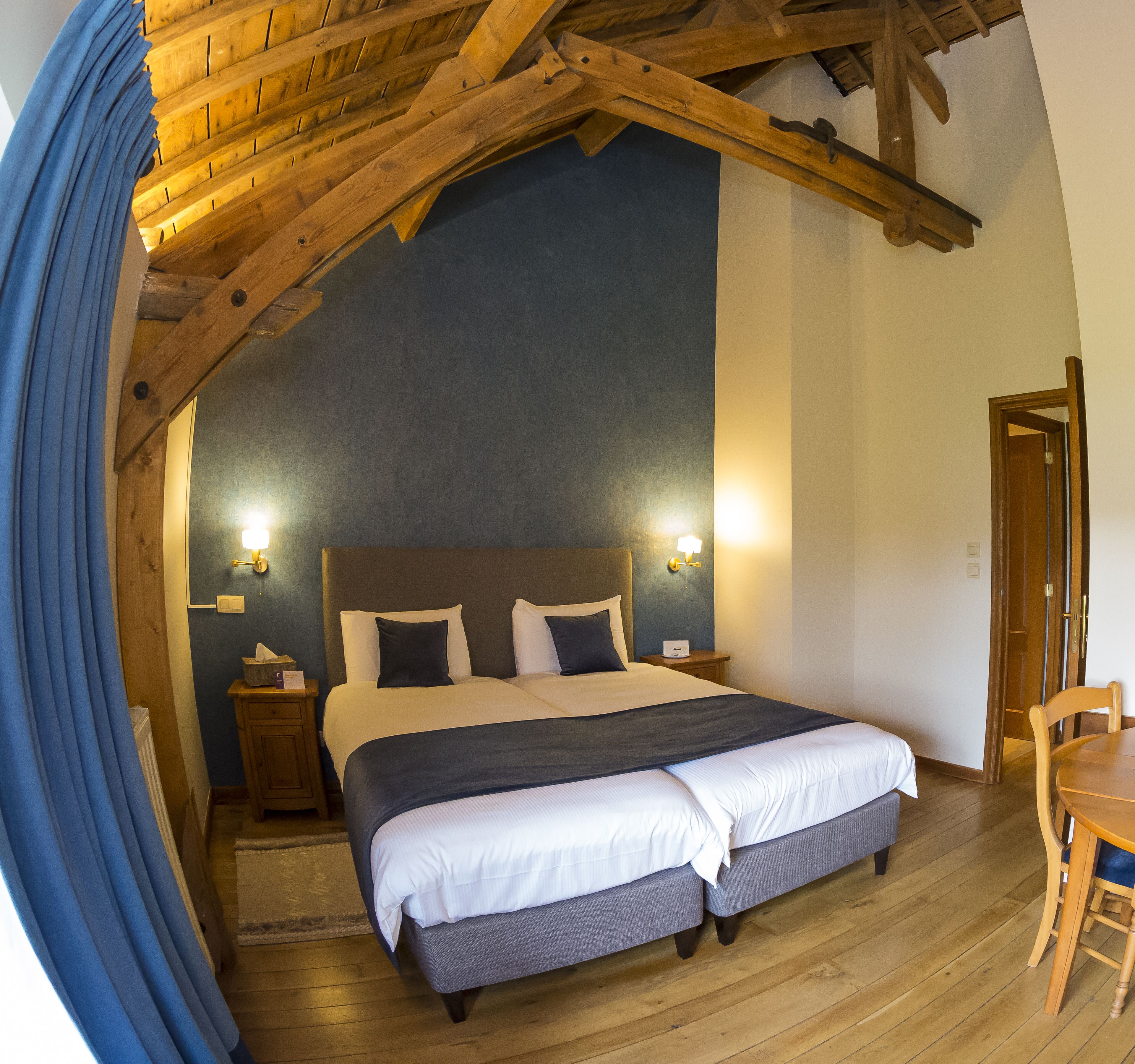 A bedroom with a large bed, wooden ceiling beams, a nightstand with a lamp, and a wooden table.