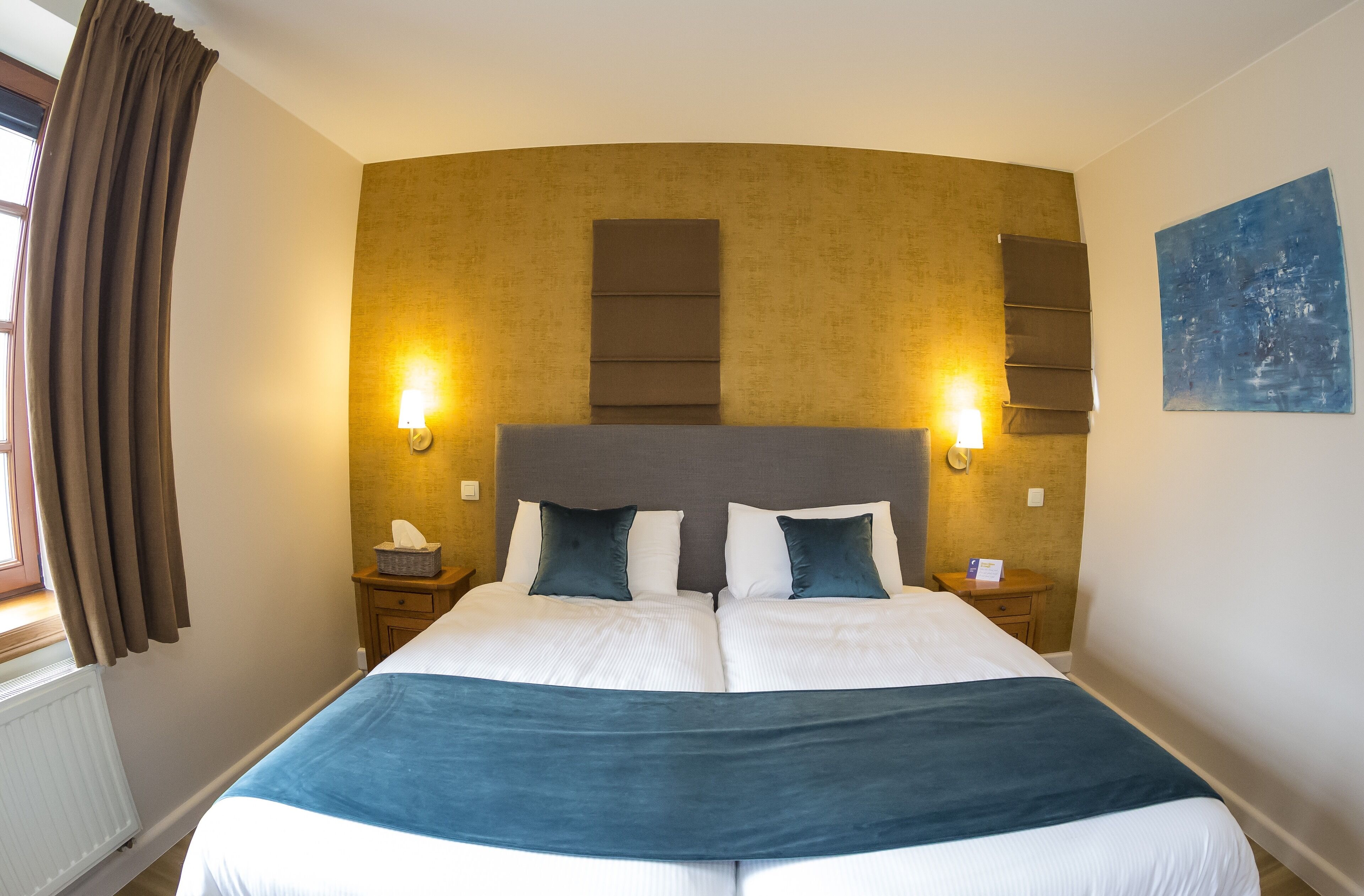A double bed with a blue throw blanket, two bedside tables with lamps, a headboard with vertical panels, a window with curtains, and a blue abstract painting on the wall.