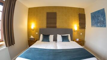 A double bed with a blue throw blanket, two bedside tables with lamps, a headboard with vertical panels, a window with curtains, and a blue abstract painting on the wall.